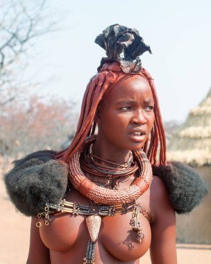 Otjikandero Himba Köyü, Namibya - 8 Eylül 2017: Himba Çamurlu kil evlerde geleneksel saç stiline, yüzüklere ve kolyeye sahip güzel Himba kadını