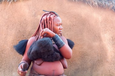 Otjikandero Himba Yetimhane Köyü, Namibya - 8 Eylül 2017: Geleneksel kızıl saçlı Himba kadını saçını tarıyor, saçlarını fırçayla tarıyor, kil çatılı arka planı var.
