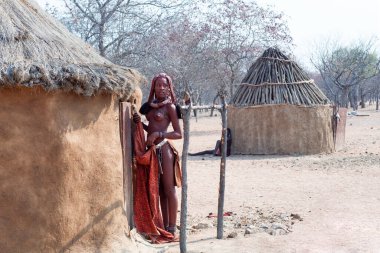 Otjikandero Himba Yetimhanesi Köyü, Namibya - 8 Eylül 2017: Himba kızı köydeki geleneksel toprak ve samandan yapılmış evin kapısında duruyor