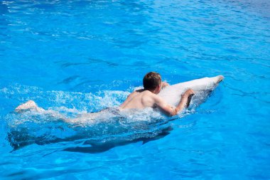 Genç bir adam yunusa biniyor, çocuk yunuslarla birlikte mavi sularda yüzüyor su havuzunda, denizde, okyanusta, yunusta bir adamı kurtarıyor.