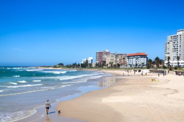 Adam sahil boyunca bir köpekle yürüyor, mavi deniz, gökyüzü, binalar ve otellerle.