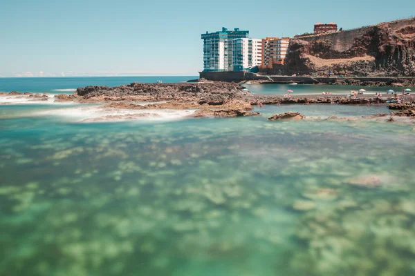 Gün batımında gökyüzünün altında binalarla çevrili şeffaf bir deniz manzarası.