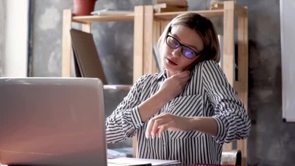 사무실에 앉아 있는 한 젊고 아름다운 소녀가 스마트폰으로 전화를 받습니다. 안경을 쓴 여자 가전 화로 정보를 받기 위해 노트에 펜으로 자료를 쓴다 — 비디오