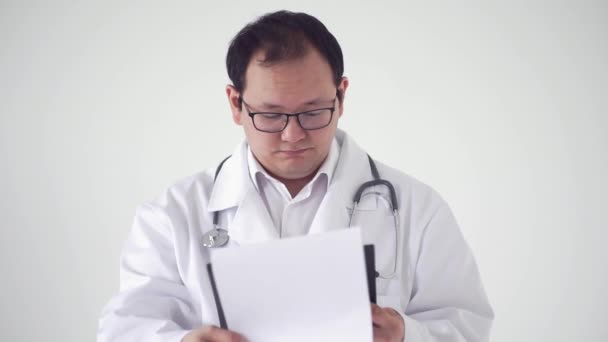 Joven médico asiático, en una capa profesional blanca con un estetoscopio, trabaja con una historia clínica y no entiende lo que está escrito, sobre un fondo blanco — Vídeos de Stock