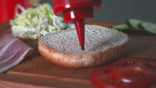 Kochen eines Burgers zu Hause. Die Hälfte des Hamburger-Brötchens ist mit roter Soße und Ketchup gefüllt. Im Rahmen von Gemüse, Tomaten, Salat, Schinken, Gurken — Stockvideo