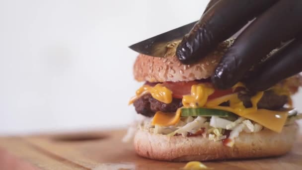 Kuchaři v černých gumových rukavicích rozřízli Burger velkým ostrým nožem v kuchyni. Zblízka. Vynikající jídlo. Šťavnaté chutné Burger na stole — Stock video
