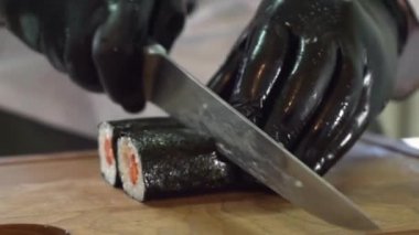 Siyah eldivenli erkek şef, mutfaktaki suşi rulolarını kesiyor. Nori pilav ve somonla servis için hazır.