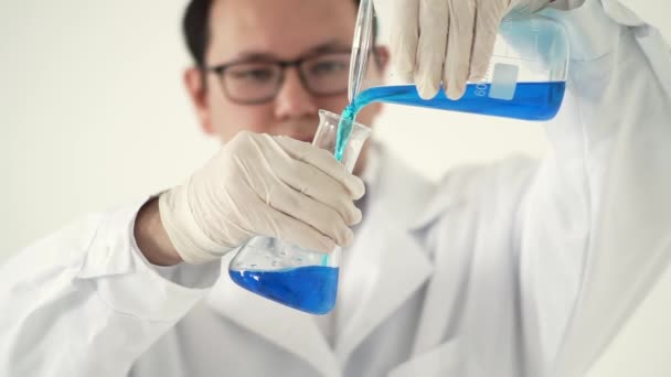 Cientista asiático em um casaco branco e luvas derrama de um grande frasco e um menor. Realiza uma experiência em um líquido azul em um laboratório — Vídeo de Stock