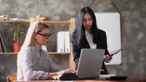 Deux belles jeunes filles créatives dans le bureau. Une femme s'assoit à une table et travaille sur un ordinateur portable, mène une conversation avec une femme en tant que chef de projet. Bureau avec des collègues sympathiques trouver — Video