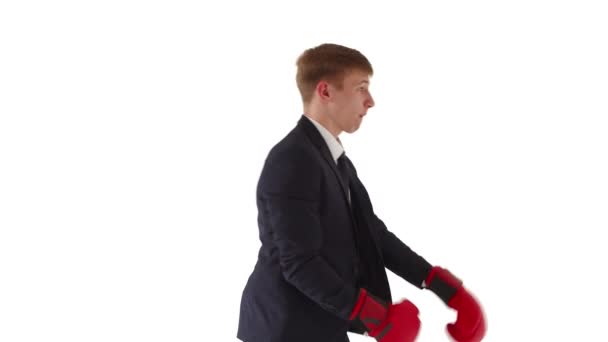 Um jovem e agressivo trabalhador de escritório se desdobra em perfil e caixa em um terno clássico e luvas de boxe vermelhas, desdobra o rosto na câmera e pede batalha. O conceito de resolver problemas de negócios — Vídeo de Stock