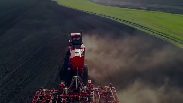 Egy traktoron dolgozó farmer felkészíti a földet a vetésre egy szántott mezőn. Magokat ültetek poros földbe. Tavaszi mezőgazdasági munka. Légi felvétel, egy drón repül előre a traktor felett. — Stock videók