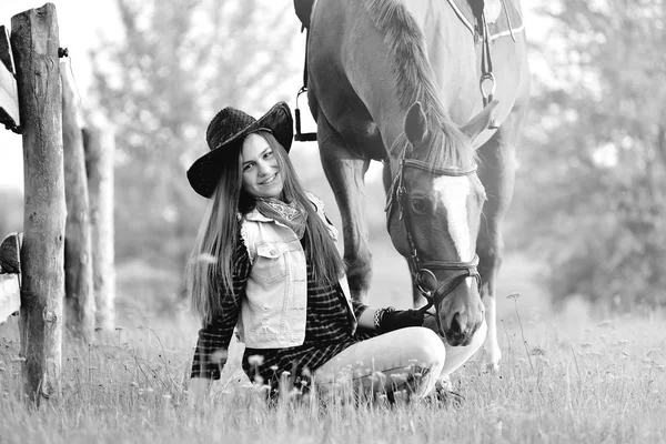 cowgirl in a hat sitting near a horse in a field