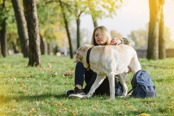 美丽的金发女郎坐在草地上的公园和她的狗拉布拉多 — 图库照片