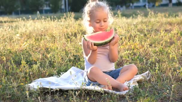 Una linda rubiecita está sentada en el parque sobre la hierba y comiendo alegremente una sandía . — Vídeos de Stock