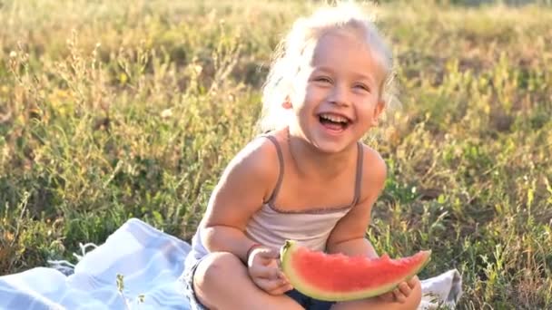 Een schattige kleine blonde is in het park op het gras zitten en vrolijk eten een watermeloen. — Stockvideo