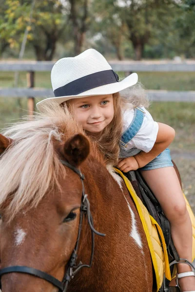 A cute little blonde girl is sitting on a pony in autumn. — Stock Photo, Image
