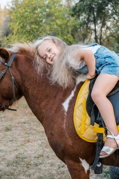 A cute little blonde girl is sitting on a pony in autumn. — Stock Photo, Image