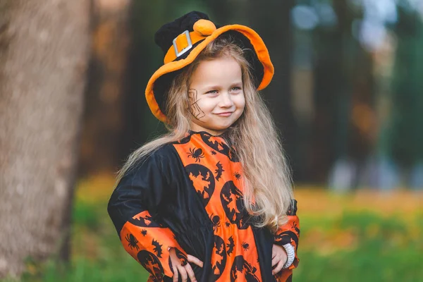 little girl in witch costume stands in the park in the fall on Halloween holiday.