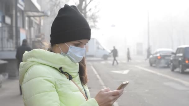 Молода жінка стоїть на міській вулиці в густому смозі в медичній масці і розмовляє по телефону . — стокове відео