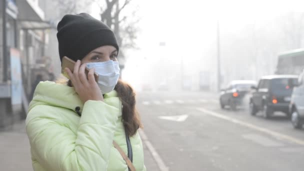 Uma jovem mulher está de pé em uma rua da cidade em densa névoa em uma máscara médica e falando ao telefone . — Vídeo de Stock