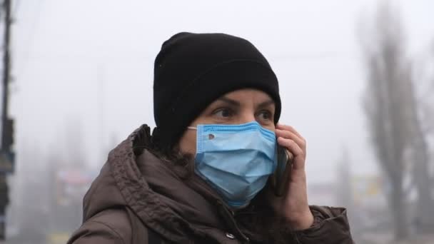 A young woman is standing on a city street in dense smog in a medical mask and talking on the phone. — Stock Video