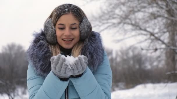 Beautiful young woman holding snow in her hands and blowing on it — Stock Video
