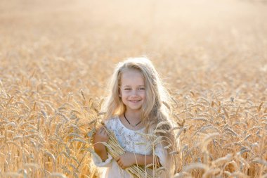 Küçük sarışın bir kız yazın elinde dikenlerle buğday tarlasında duruyor ve gülümsüyor..