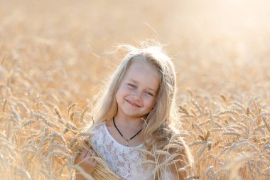Küçük sarışın bir kız yazın elinde dikenlerle buğday tarlasında duruyor ve gülümsüyor..