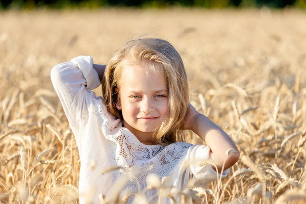 Klein blond meisje staat in een tarweveld in de zomer en glimlacht — Stockfoto