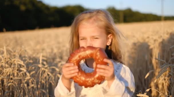 Niña divirtiéndose y comiendo un bollo en un campo de trigo en verano . — Vídeos de Stock