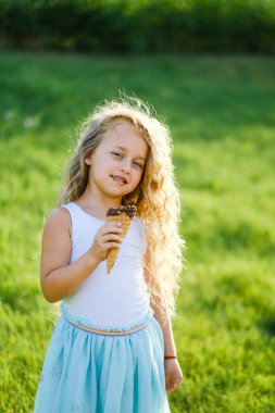 Uzun saçlı küçük sarışın kız yaz parkında dondurma yerken eğleniyor. Bir kız yaz parkında yürüyor ve dondurma yiyor. Mutlu çocukluk ve mutlu yaz.