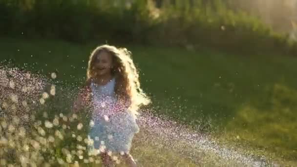 小さな女の子は緑の芝生で楽しんでいます 夏には水の流れが注ぎ込まれます 芝生の上を流れる水の下で暑い夏の冷却 — ストック動画