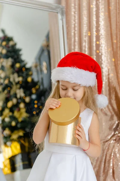 Little cute girl in Santa hat opens a Christmas present. — Stock Photo, Image