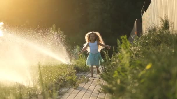 少女は夏に芝生に水をやるために水のジェットを通って走ります 子供は冷えていて 暑い夏の日に水の流れの近くで楽しんでいます — ストック動画