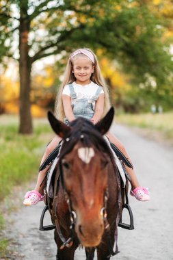 Uzun saçlı küçük sarışın kız sonbaharda günbatımında parkta ata biner. Sonbahar ata binme. Bir kızla bir atın arkadaşlığı..