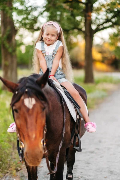 Malá blondýnka s dlouhými vlasy jezdí na koni v parku při západu slunce na podzim. Podzimní jízda na koni. Přátelství dívky a koně. — Stock fotografie