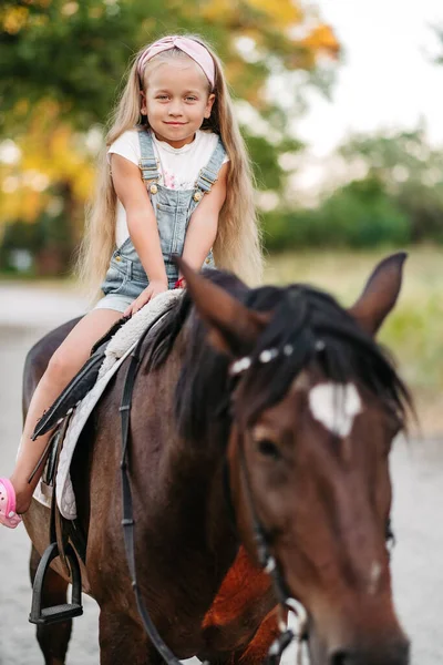 Petite blonde aux cheveux longs monte à cheval dans le parc au coucher du soleil en automne. Promenade à cheval d'automne. Amitié d'une fille et d'un cheval. — Photo