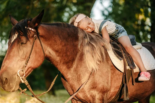 Mała blondynka z długimi włosami kłamie i przytula się na koniu w parku o zachodzie słońca jesienią. Jesienna przejażdżka konna. Przyjaźń dziewczyny i konia. — Zdjęcie stockowe