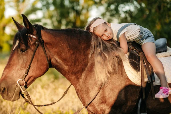 A little blonde girl with long hair lies and hugs astride a horse in the park at sunset in autumn. Autumn horse ride. Friendship of a girl and a horse. — Stock Photo, Image