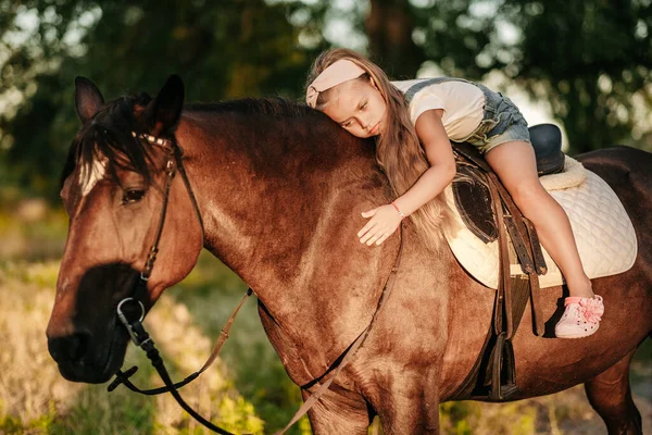 Malá blondýnka s dlouhými vlasy leží a objímá se na koni v parku při západu slunce na podzim. Podzimní jízda na koni. Přátelství dívky a koně. — Stock fotografie