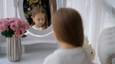 Küçük kız Noel için hazırlanıyor. Küçük sarışın aynanın önünde oturur ve kendine bakar. Noel Arifesi.