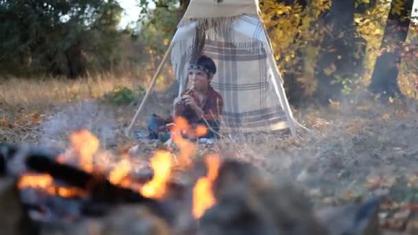 一个身穿美洲原住民服装的小男孩坐在维格瓦姆旁边弹奏长笛 在火边和印地安人玩 头上顶着羽毛 美洲原住民遗产日 — 图库视频影像