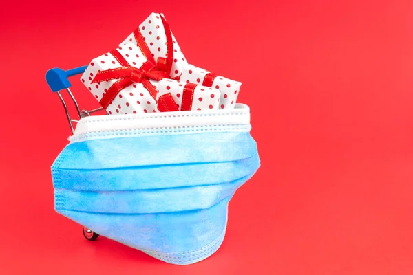 Kırmızı arka planda, koruyucu bir maskeyle fotokopi alanı olan bir alışveriş arabası. Virüs esnasındaki bayram hediyesi konsepti..