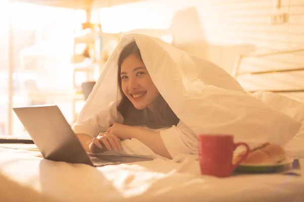 Pretty asian woman working on laptop computer. Concentrated female lying in bed at home writing and using notebook keyboard touchpad. Freelance lifestyle and work from home