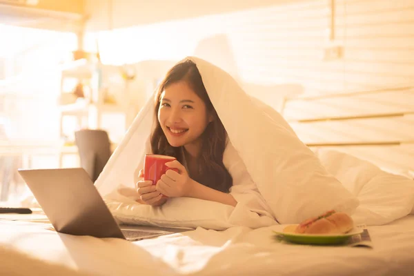 Asian woman working on laptop computer. Relax female lying in bed at home and hold a cup of coffee. Freelance lifestyle and work from home