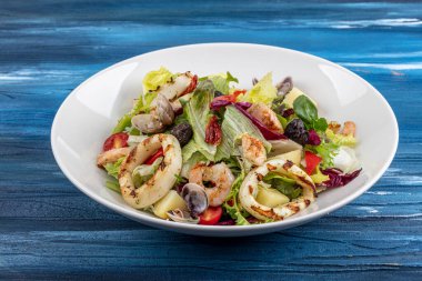Deniz mahsullü salata. Lezzetli taze karides salatası, kalamar, karides, midye ve karışık marul yaprakları, mavi ahşap masadaki beyaz tabakta roka, üst manzara..