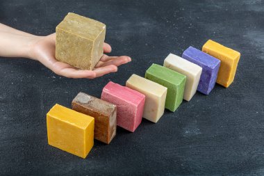 Doğal zeytinyağı sabunu tutan bir kadın. Siyah ahşap arka planda birçok farklı el yapımı sabun çubukları.