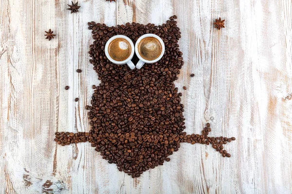 Coffee beans on wooden background form the silhouette of an owl and two cups of coffee form their eyes - concept with coffee and night owls.