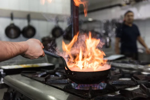 Chef Cooking With Fire In Frying Pan. Professional chef in a commercial kitchen cooking flambe style. Chef frying food in flaming pan on gas hob in commercial kitchen.