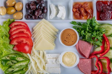 Gri ahşap masada geleneksel Türk kahvaltı tabağı, üst görünüm: pogaca hamur işleri, sebzeler, peynir, zeytin ve helal hindi salamı, seçici odak noktası; metin için kopyalama alanı.